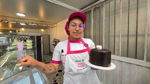 Pastelera en uniforme rosa sostiene un plato blanco con una torta de chocolate decorada con crema y una cereza. 