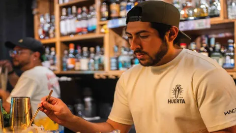 Bartender de Huerta Coctelería decorando un cóctel en la barra con botellas de licor de fondo.