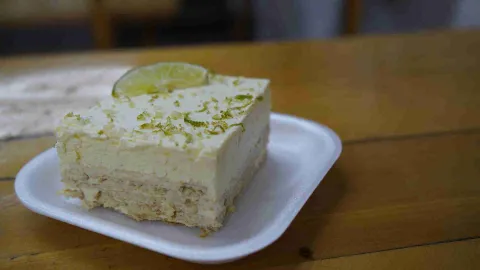 Porción cuadrada de postre de limón con ralladura cítrica y una rodaja de lima servida sobre un plato desechable. 