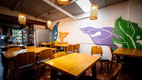 Comedor con mesas de madera, lámparas tejidas de mimbre y pared pintada con ilustración de hojas verdes y moradas.