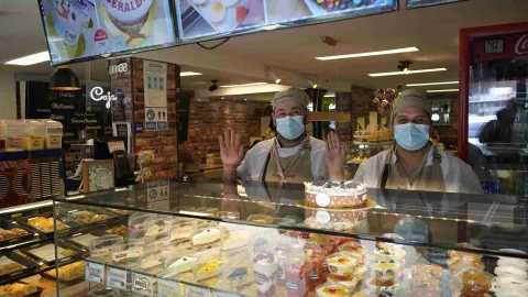 Dos empleados con tapabocas saludando detrás de la vitrina exhibidora con postres y tortas variadas. 