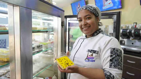 Una chef sonriente muestra un postre de maracuyá envasado, junto a una vitrina refrigerada llena de otros dulces.