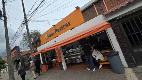 Entrada del local Solo Postres con toldo naranja, personas conversando en mesas de madera y peatones transitando.