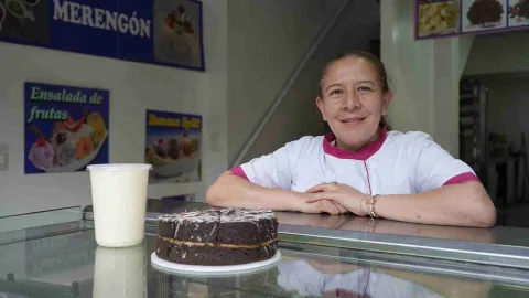  Repostera apoya sus brazos en el mostrador de vidrio junto a un pastel de chocolate y un vaso de bebida cremosa.