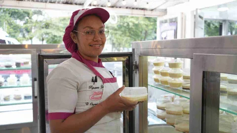Pastelera sonríe mientras saca un recipiente individual con postre de una vitrina refrigerada de acero. 