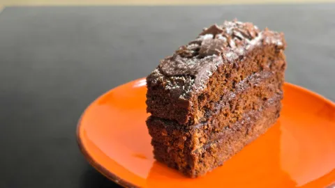 Porción de pastel de chocolate de varias capas con cobertura y trozos de chocolate sobre plato naranja
