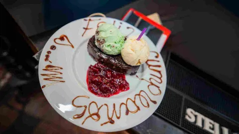 Plato de brownie con helado de menta y vainilla, mermelada de frutos rojos y dedicatoria decorativa en el borde.