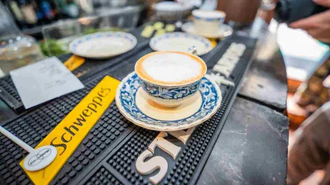  Taza de café con leche espumosa servida en porcelana decorada sobre un tapete de bar de marca Schweppes.
