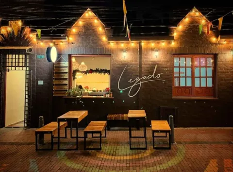 Fachada nocturna de ladrillo con luces cálidas, letrero luminoso de Legado y mesas en la terraza exterior. 