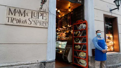 Plano medio de la entrada con placa de Mamá Lupe Tradición Santafereña, menú en marco rojo y empleado junto al portal. 