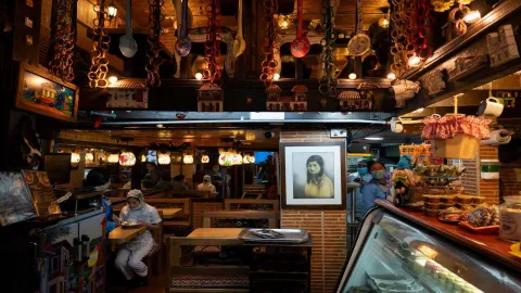 Interior del restaurante con mesas de madera, cuadro en pared de ladrillo, vitrina de postres y adornos colgantes.