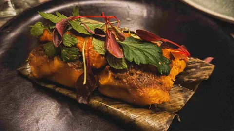 Preparación de sabores intensos, servida sobre hoja y acompañada de vegetales asados, hierbas frescas y delicados brotes.