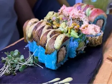 Sushi roll de arroz azul con relleno de pescado y vegetales, cubierto con salsas, semillas y toppings, servido sobre una bandeja negra.