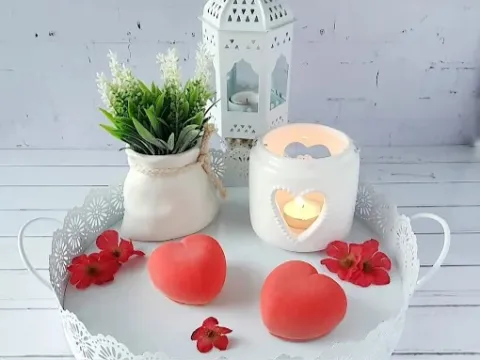 Bandeja decorativa blanca con dos postres en forma de corazón rojo, flores naturales y velas encendidas, acompañados de una planta ornamental en un ambiente romántico.