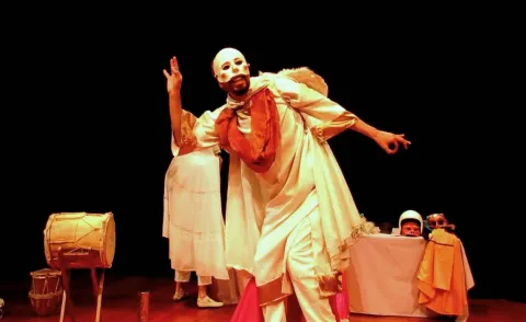 Un artista con máscara blanca y vestuario tradicional protagoniza una escena teatral llena de expresión, música y simbolismo, acompañado de tambores y otros elementos escénicos.