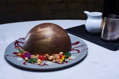 Postre de chocolate en forma de esfera, decorado con frutos, flores comestibles y salsa de frutos rojos.