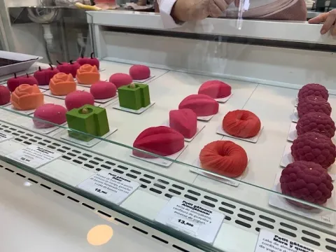 Vitrina de Helga Blaha con una selección de postres de autor en formas geométricas y diseños creativos, en tonos rosados, rojos, naranjas y verdes, exhibidos cuidadosamente sobre bandejas individuales