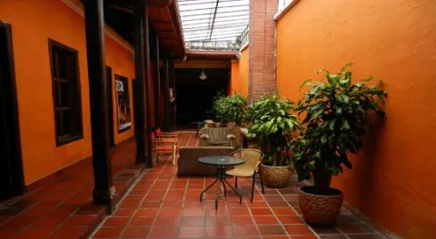 Patio interior de arquitectura colonial, con paredes naranjas, corredores de madera, plantas ornamentales y mesas que crean un ambiente acogedor y tranquilo.
