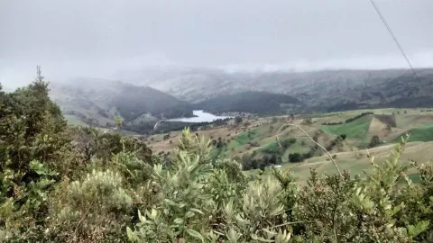 Vista panorámica de montañas verdes, vegetación nativa y terrenos rurales, con una laguna al fondo bajo un cielo nublado.