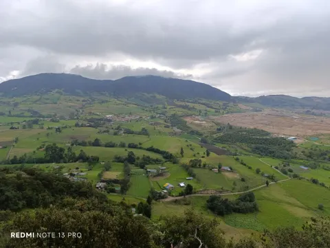  Vista panorámica del paisaje rural de Ciudad Bolívar, con campos verdes, pequeñas viviendas y montañas bajo un cielo nublado.
