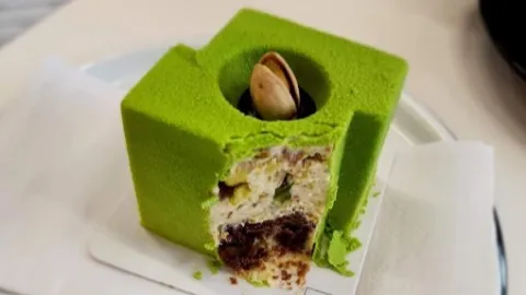 Postre de autor de color verde intenso, con forma cúbica, relleno de varias capas de crema y bizcocho, y decorado con un fruto seco en el centro.