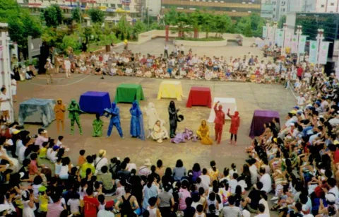  Público reunido en una plaza para disfrutar de una presentación de teatro callejero, con artistas vestidos de colores y una puesta en escena al aire libre.