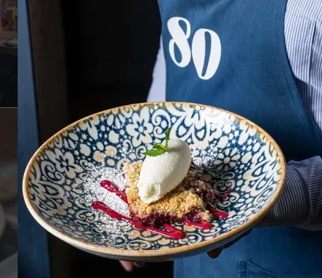 Postre artesanal de 80 Sillas, servido en un plato decorativo, con crema, frutos rojos y crumble.
