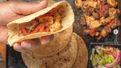 Mano sosteniendo una tortilla de maíz rellena de pollo salteado y pimientos rojos sobre una plancha con fajitas.