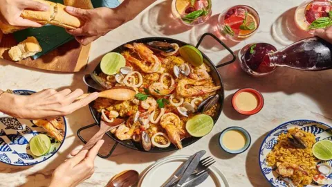 Vista superior de una paella marinera con camarones, mejillones y calamares rodeada de comensales y bebidas.