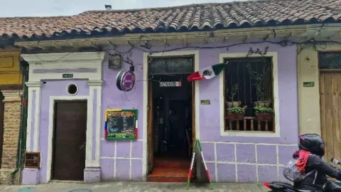 Entrada a una casa colonial pintada de lila con zócalos morados, bandera mexicana y letrero comercial de tacos.