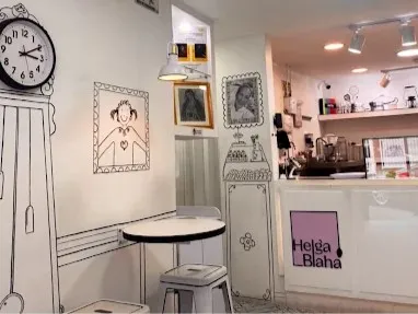 Interior de Helga Blaha, con mesas pequeñas, ilustraciones decorativas en las paredes y una barra de atención donde se exhiben productos de pastelería, en un ambiente acogedor y creativo.