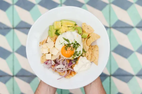 Plato servido en un tazón blanco con nachos, aguacate, huevo pochado, queso en cubos, cebolla morada y crema.