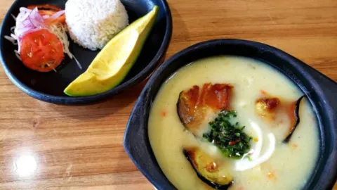 Sopa tradicional servida en cazuela de barro junto a un plato con arroz blanco, ensalada y tajada de aguacate. 