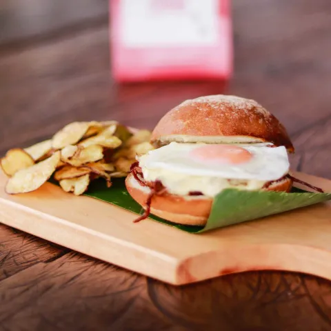 Hamburguesa con huevo frito, queso y tocineta, acompañada de chips de papa sobre una tabla de madera.