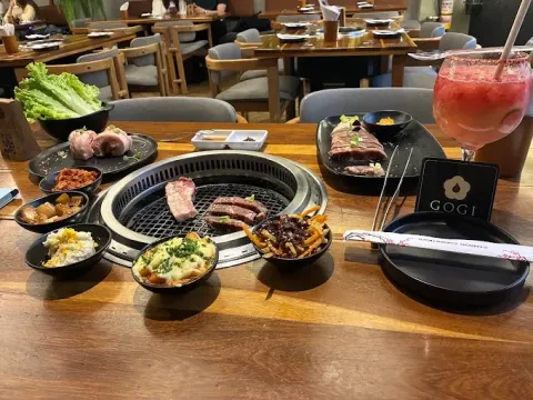 Mesa de barbacoa coreana con parrilla central, cortes de carne, acompañamientos, salsas y una bebida refrescante.