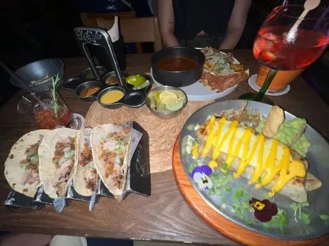Mesa con tacos, platos de comida mexicana, salsas, guacamole y bebidas, en un ambiente de celebración.