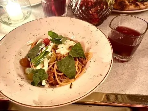 Pasta servida con tomates, queso fresco y hojas verdes, acompañada de una bebida.