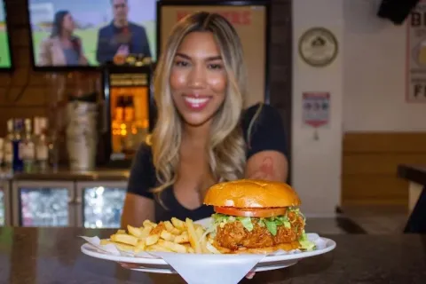 Mujer joven muestra un plato de Hamburguesa acompañada de papas fritas, servida en mesa de Hooters.