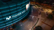 The Movistar Arena facade at night, with the stage illuminated in blue and a sign in white neon lettering. In front is a small plaza made of brick, concrete, and vegetation.