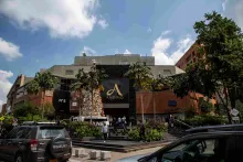 Facade of a building with at least three stories, made of brick and concrete, featuring a large, gold letter A and the word ""Andino."" In front, there are stairs with people and a few cars nearby.