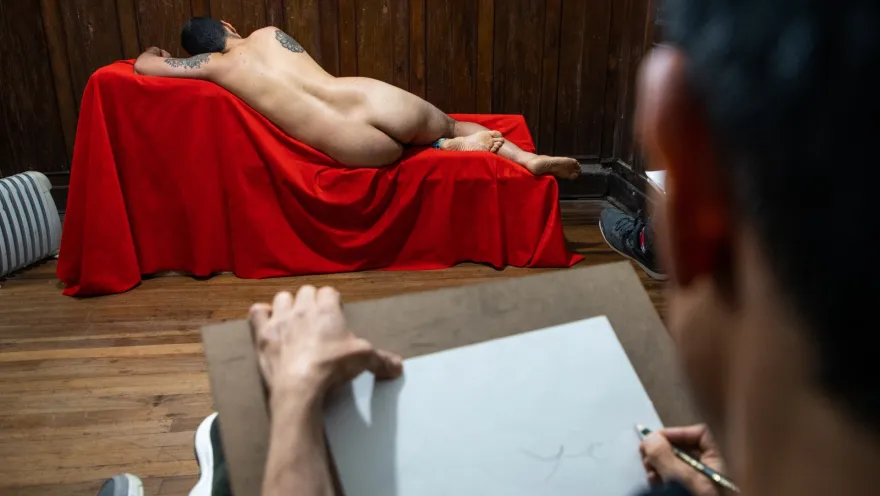 Chucuy Club con hombre jóven desnudo de espalda y sobre una silla cubierta con una tela roja es dibujado por otro hombre que hace el gráfico en un papel blanco.