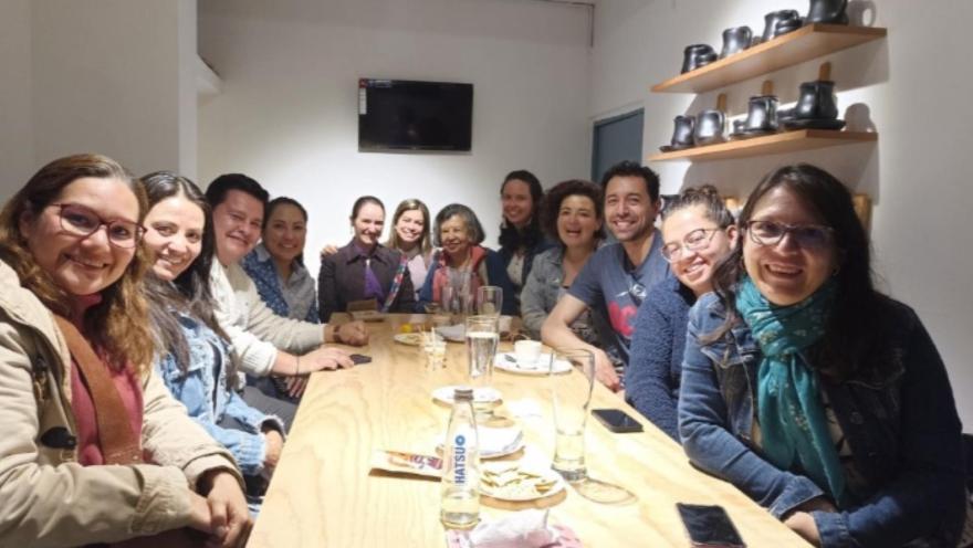Grupo de personas en un salón, mayoría mujeres jovenes y adultas ríen mirando al frente. Al lado en una repisa en la pared hay unos tazones negros de chocolate.