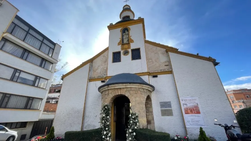 Parroquia Santa Monica fachada en tarde soleada con cielo azul, puerta decorada con flores. Al lado izquiero hay un edificio de apartamentos.