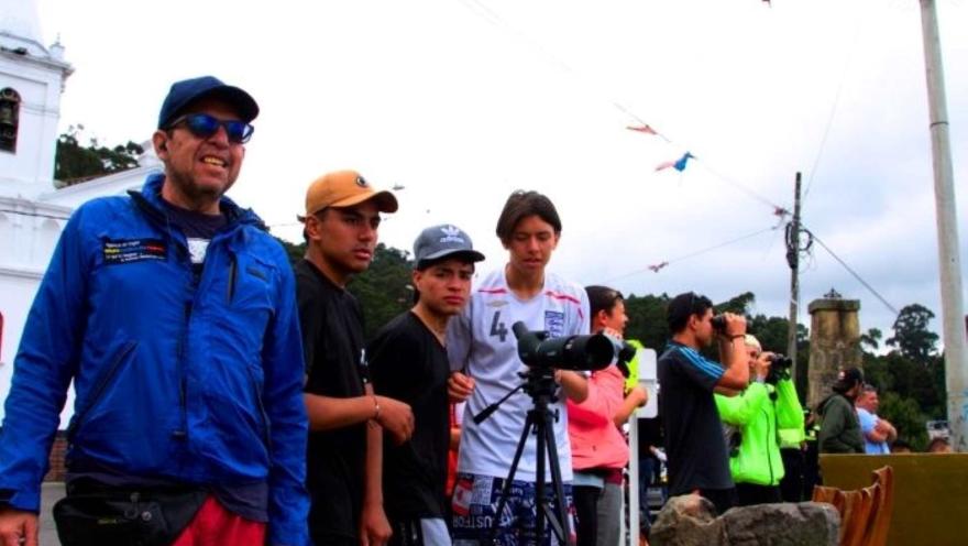 Grupo de personas jóvenes junto con un guía hombre, observando un mirador. Todos con ropa deportiva, algunos hombres llevan gorra, otros binoculos.