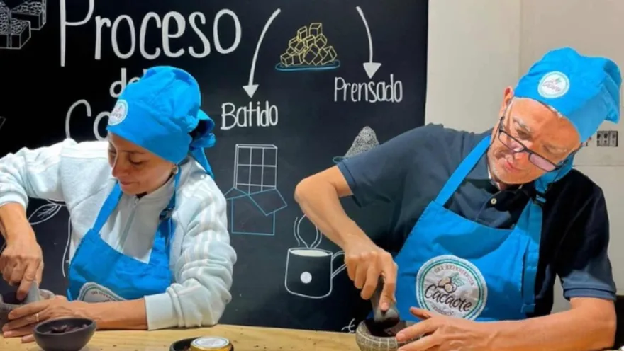 Un hombre y una mujer adultos con delantales y gorros azules muelen cacao en sendos morteros. Detás hay un aviso que señala el recorrido de la transformación del chocolate