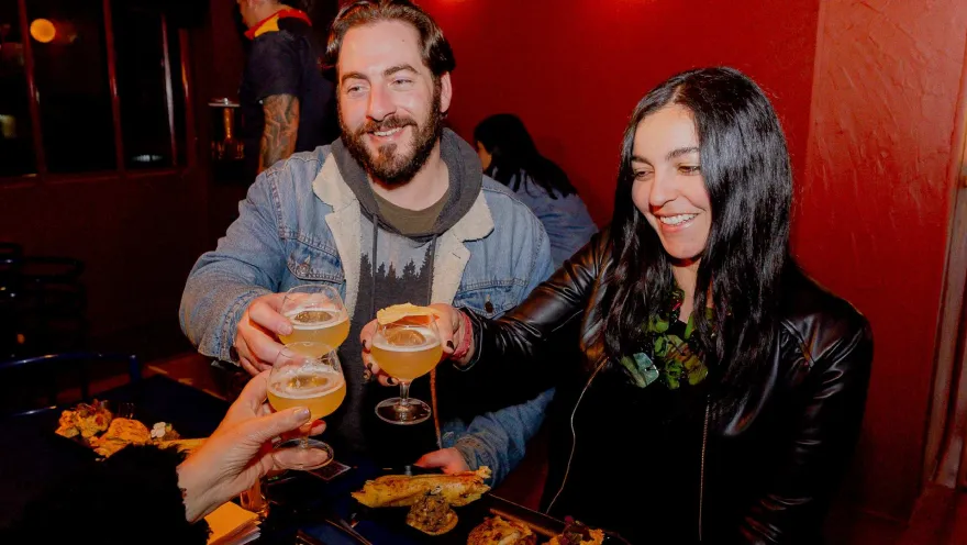 Un hombre y una mujer jóvenes brindan con copas con licor alrededor de una mesa con cómida típica colombiana. como empanadas, arepas y papas criollas.