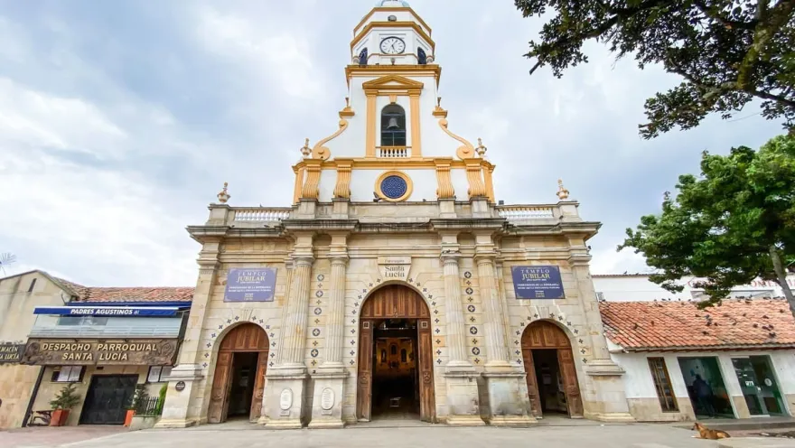  Iglesia Parroquial Santa Lucia - Chia con fachada frente con tres gigantes puertas en madera café  en contrapoicada con cielo azul y blanco. Un perro amarillo descansa al lado de una de las puertas.