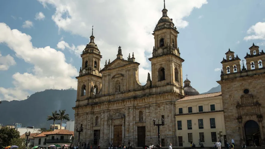 La Candelaria – Primatial Cathedral, wide shot of the entire facade, along with the Church of El Sagrario. In front, people and pigeons; in the background, the Eastern Hills under a blue sky.