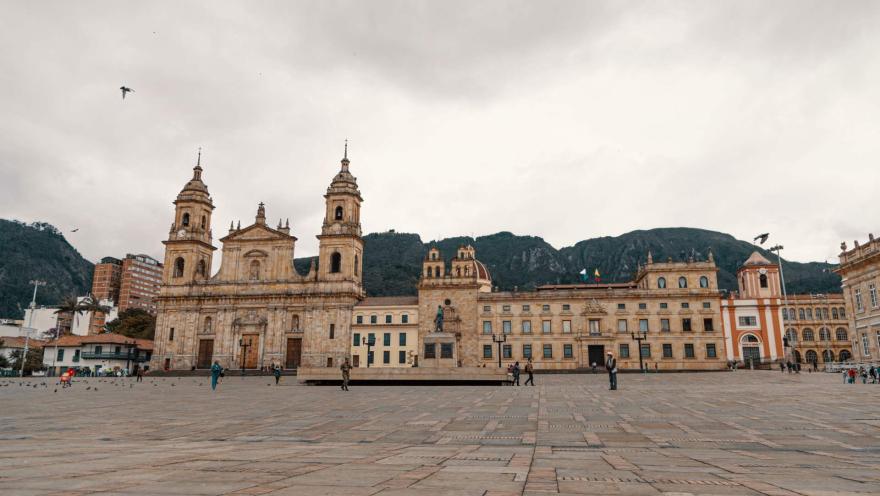 Plaza de Bolívar vista desde el occidente con algunas personas a lo lejos y de fondo la Catedral Primada y el Sagrario. Al fondo los cerros orientales y el cielo blanco con dos palomas en vuelo.