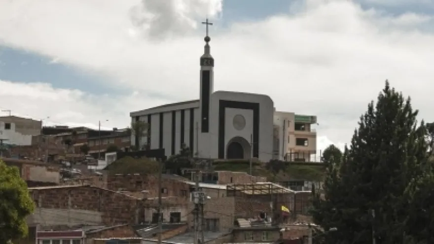 Santuario Nuestra Señora de la Peña vista de lejos en una cima detras de casas de barrio popular con cielo azul y nubes blancas. Destaca su fachada blanca con visos negros, su entrada en forma de óvalo y la torre con una punta alta que cierra con una cruz. 